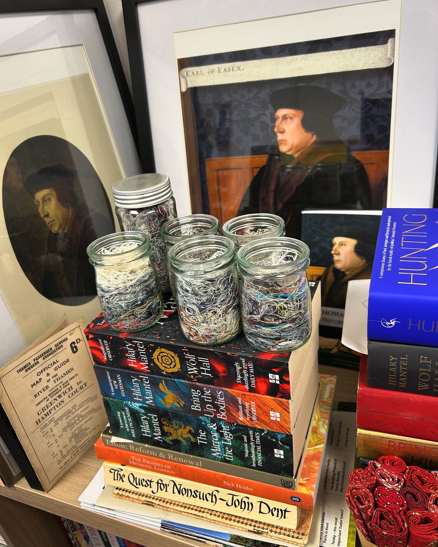 Six jars of cut thread resting on a pile of books about Thomas Cromwell and Tudor England. There are portraits of Thomas Cromwell leaning up on the wall behind the pile of books.