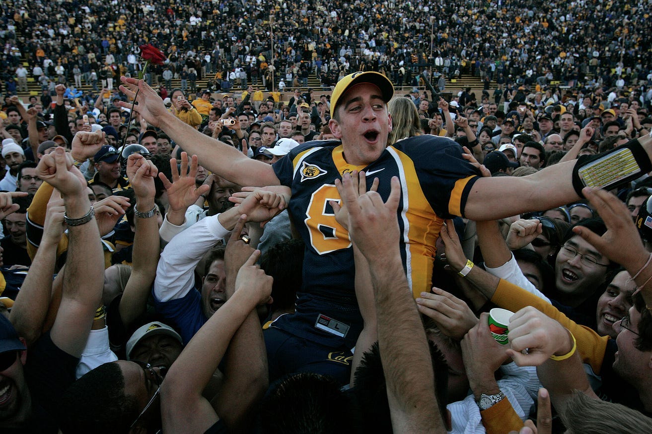 A college age Aaron Rodgers being celbrated on the field. A crowd lifts him up, his arms outstrecthed, as he holds a rose in his hand.