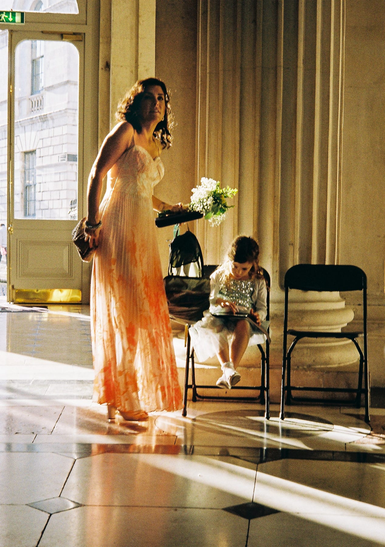 Golden light bathes a woman in a bridesmaid dress carrying a bouquet of flowers as she stand over a seated girl who is looking at a phone in a by a marble column.