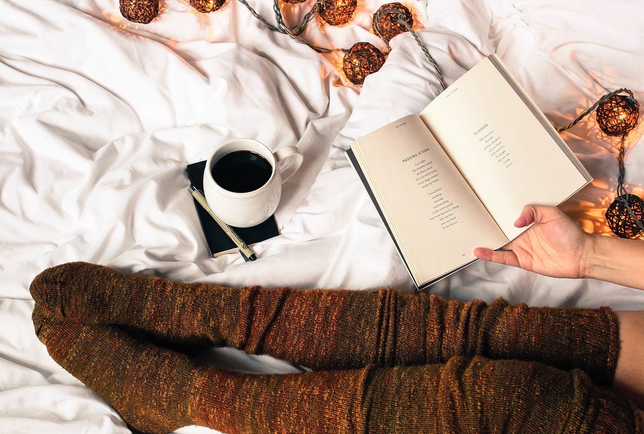 Person wear brown knitted stockings, reading poems, with a cup of tea, a note pad and pen available. Surrounded by decorative lights.