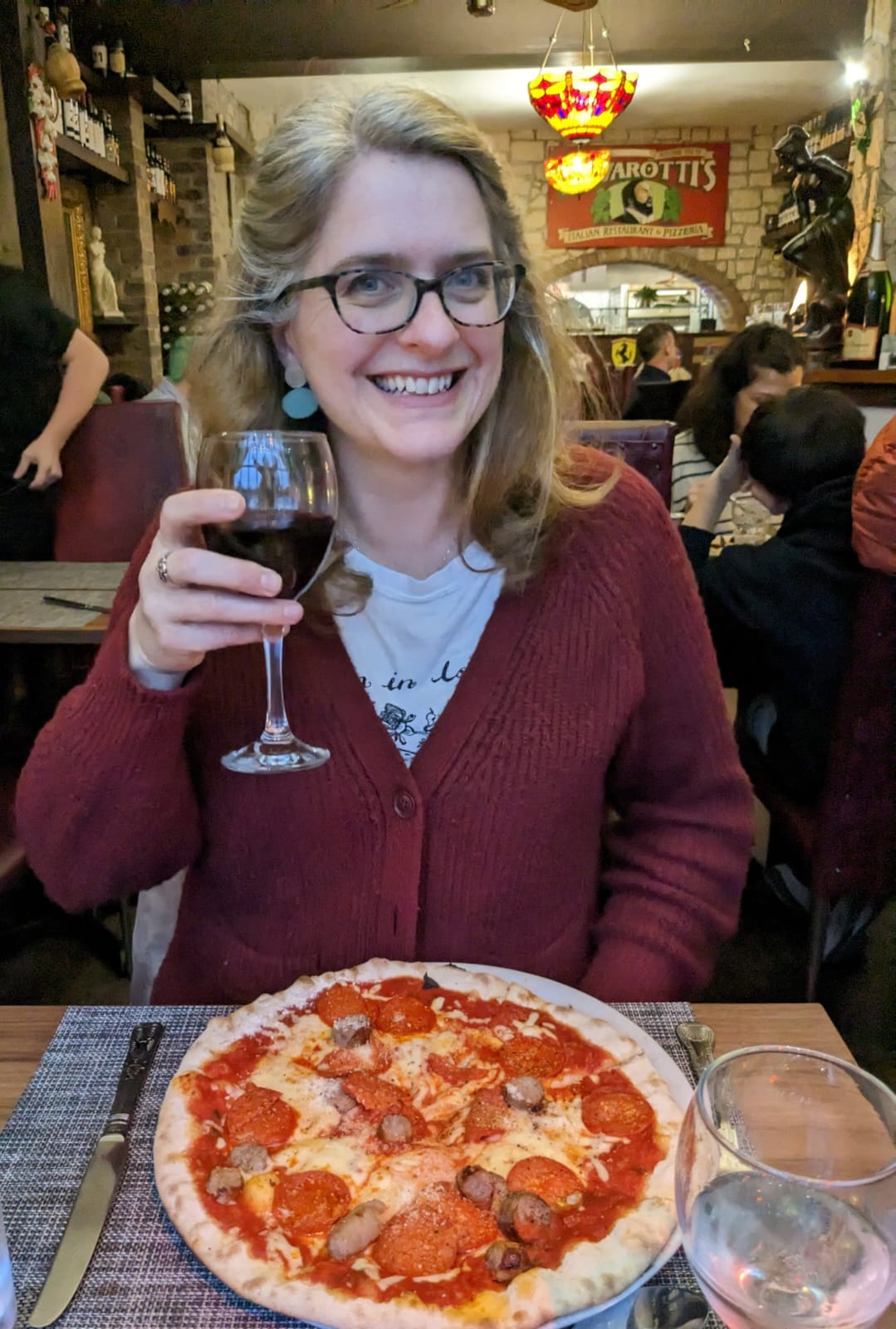 Photo shows a smiling Emma in a restaurant with a pizza in front of her, raising a glass of red wine. 