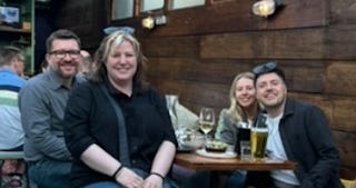 Four people, two men and two women (one of them me at the front) siting around a beer garden table. There glasses of wine and beers, and small plates of food. Everyone is smiling with sunglasses on our heads, happily posing for the photo.