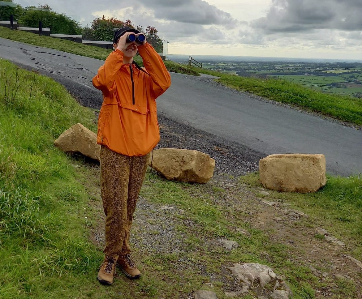 Me (Janelle) a tall woman in a bright orange top standing up. I've got binoculars to my eyes. Behind me are green fields of Lancashire in the direction of Blackpool