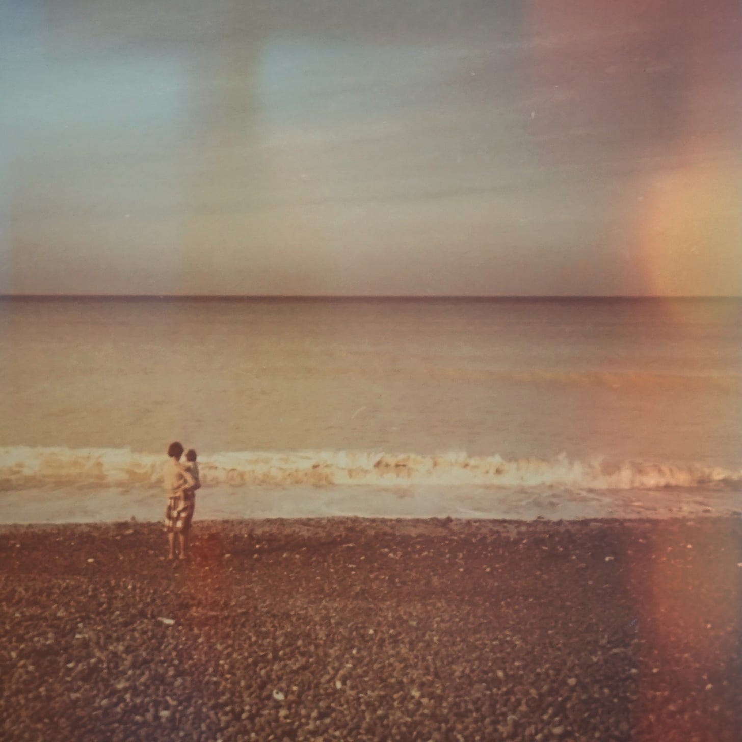 uma mulher segura uma criança na praia, olhando para o mar.