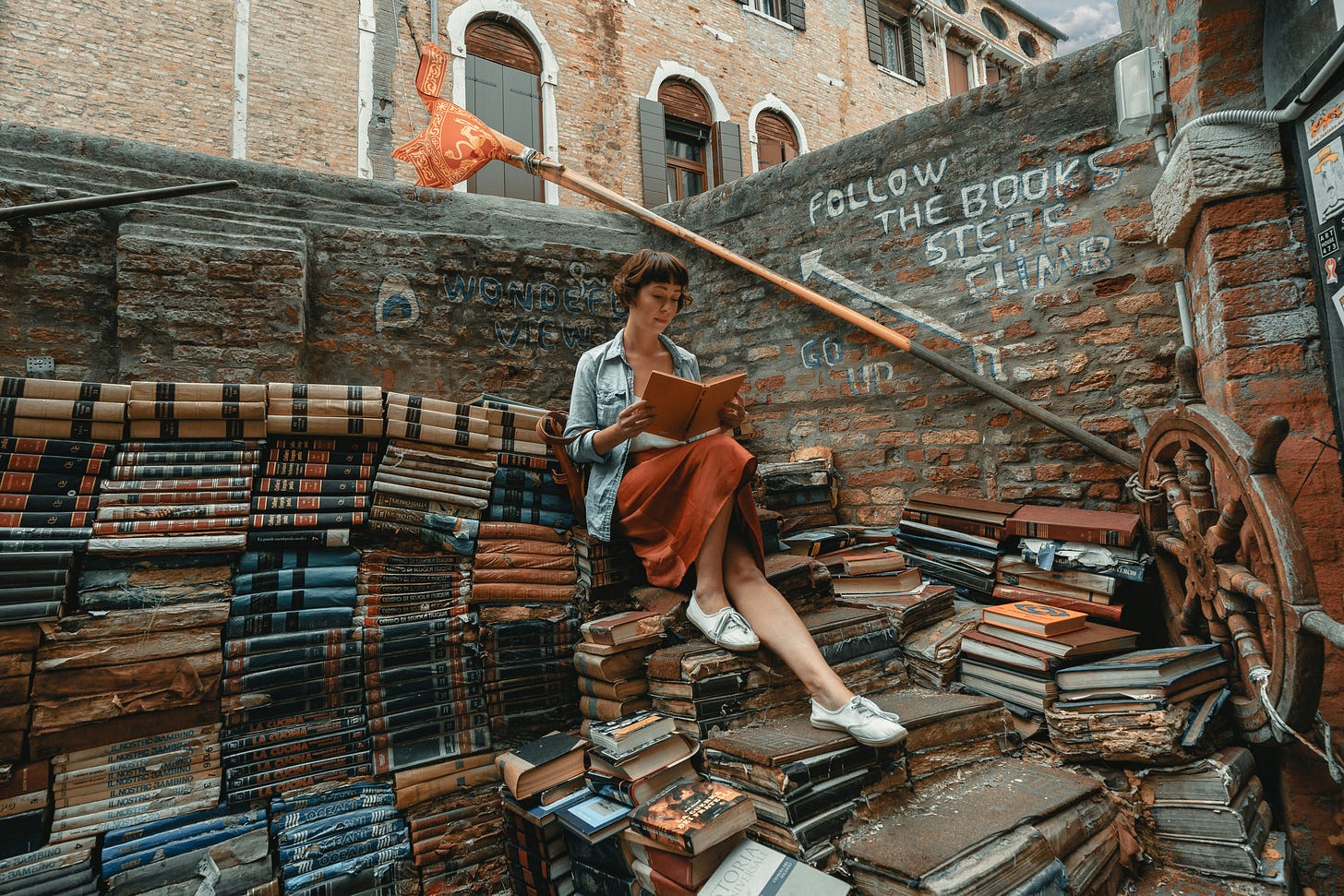 Woman reading outdoors surrounded by piles of books. A sign on a brick wall reads "Follow the books / Steps / Climb," and points up a few stairs