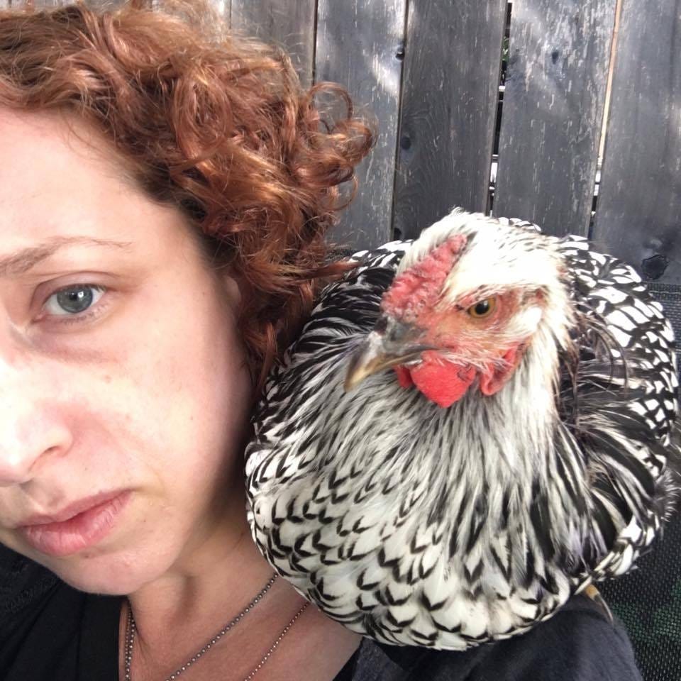A black and white chicken sits on the shoulder of a redheaded woman, Both look at the camera