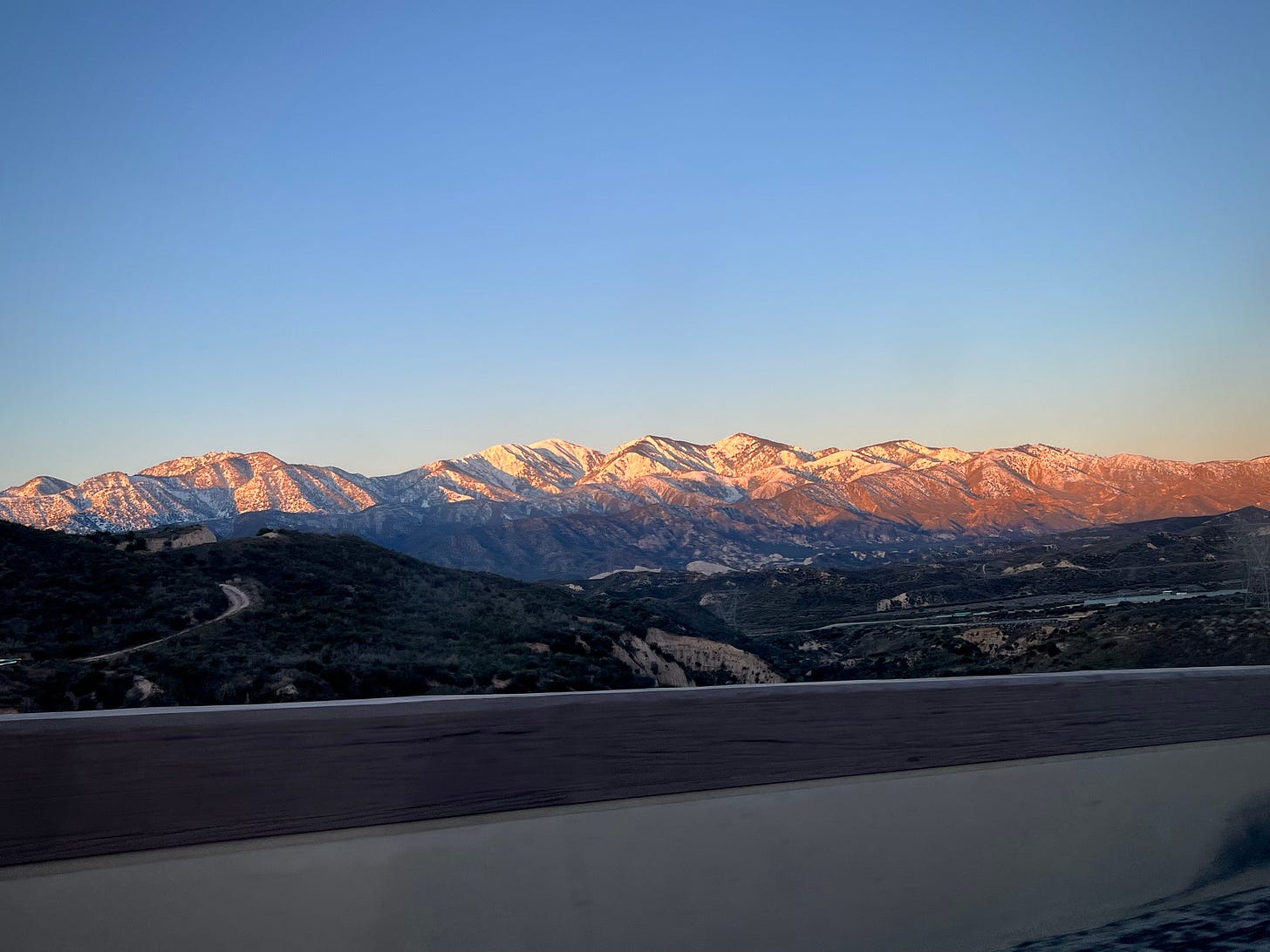 Periwinkle sky, a mountain range (peaks coated in snow), and below them hills, highways, and the side of the road we're driving on. It's sunrise, so the mountains are illuminated in gold light in the top parts.