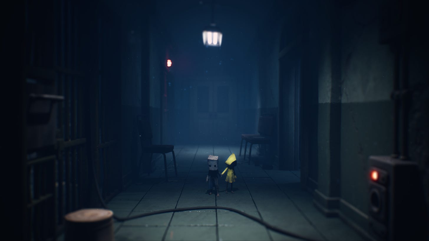 A screenshot from Little Nightmares 2, where Six and Mono are coming down a long hallway in the Hospital level. Mono, with his paper-bag head is on the left, holding hands with Six in her signature yellow raincoat on the right. They are dwarfed by the scale of their surroundings.