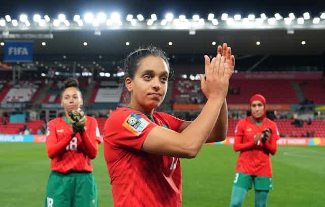 Three women on the field in a sports stadium, dressed in red tops and green shorts and leggings, one with a hijab. The woman in front raises her hands in applause.