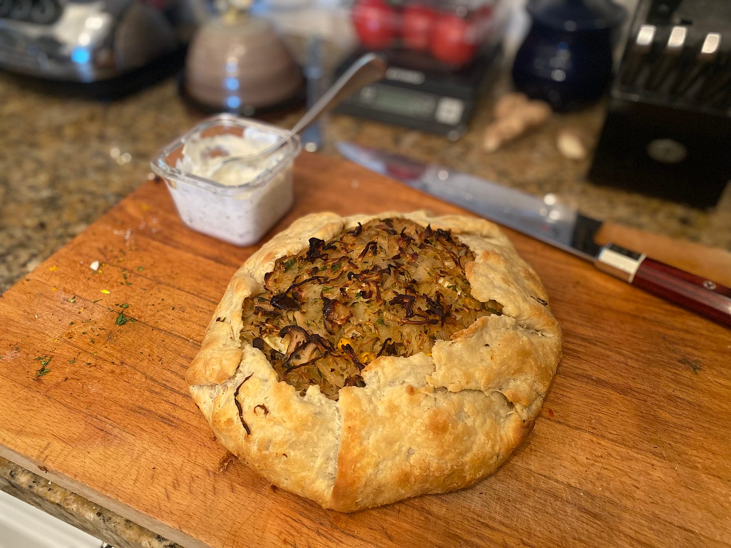 The galette described above, with browned folded edges and the vegetables in the centre brown and crisped. It's on a wood cutting board with a large chef's knife and a square container of the sour cream mix, with a spoon in it, visible in the background..