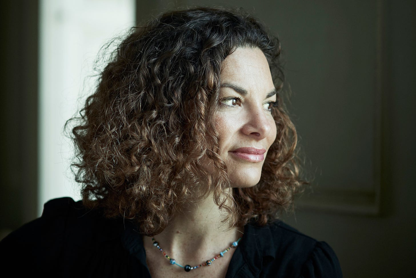 A photo of the author Gaia Vince. It is a cropped head-and-shoulders shot. Vince's head is turned to face the right of the image. She has medium-length curly brunette hair and is wearing a colourful, short necklace.