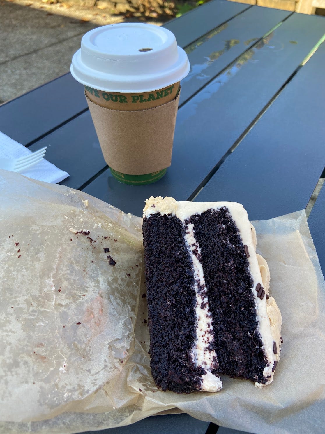 A piece of chocolate salted caramel cake and a to-go cup of coffee on a black picnic table.
