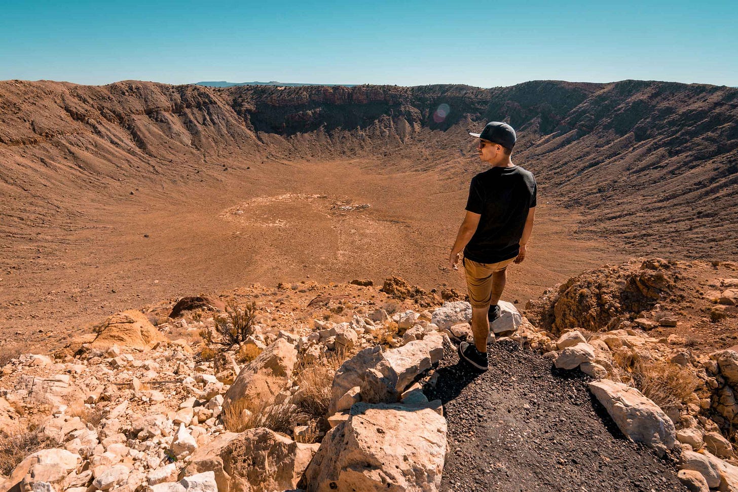 A man in baseball cap standing at the edge of a mile-wide meteor crater with desert surroundings A man in baseball cap standing at the edge of a mile-wide meteor crater with desert surroundings