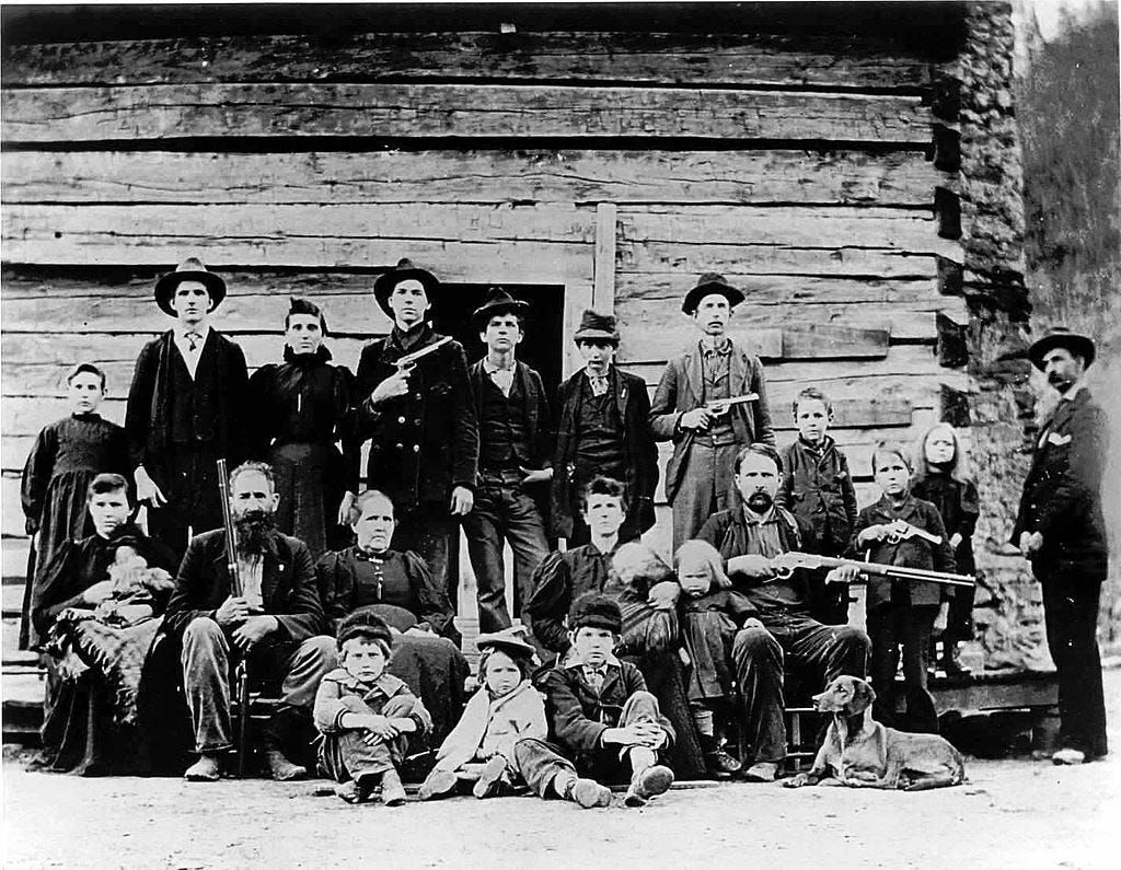 A black-and-white photo of a pioneer family posed in front of a log cabin, featuring men, women, and children armed with rifles, symbolizing the rugged lifestyle of early settlers.