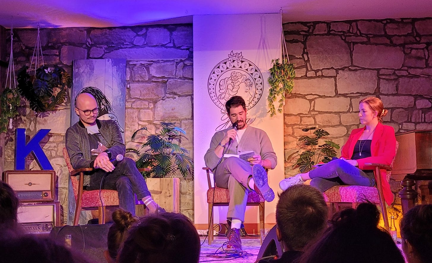 Die Autoren Daniel Stähr und Simon Sahner sitzen mit der Moderatorin Martha Routen auf der Bühne des Freiburger Kulturaggregats. Sahner liest aus dem Sachbuch „Die Sprache des Kapitalismus“ vor. (Foto: vliestext)