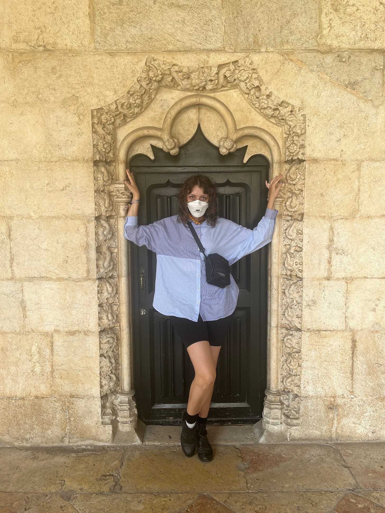 Eu fazendo pose numa portinha do Mosteiro dos Jerônimos, em Lisboa. Estou usando camisa azul claro, short preto, botas pretas e máscara aura