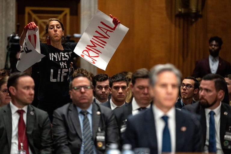 A protester wearing a shirt that reads "Invest in Life" stands and holds two signs, one of which reads "Criminal." Her hands are painted red.Text on the other can't be seen in the photo. Men are seated around her, including an out of focus Anthony Blinken in the foreground.