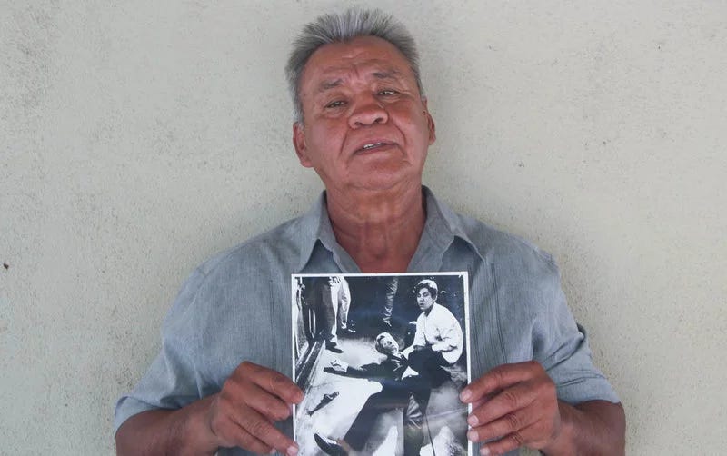 An older Mexican man stands in front of a white wall. The man is wearing a light blue shirt. In the man's hand he is holding a photograph of him as a young man with Robert F. Kennedy after he has been shot. 