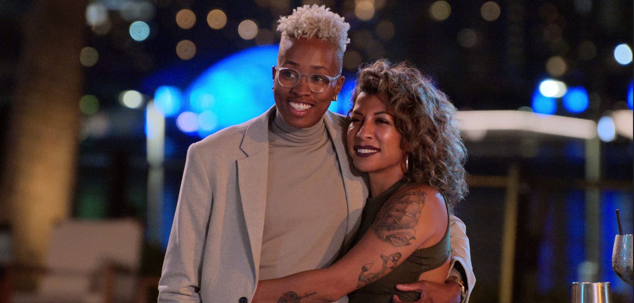 a couple: one a black woman with short blonde hair, and the other a latina woman with longer curly hair and tattoos, embrace