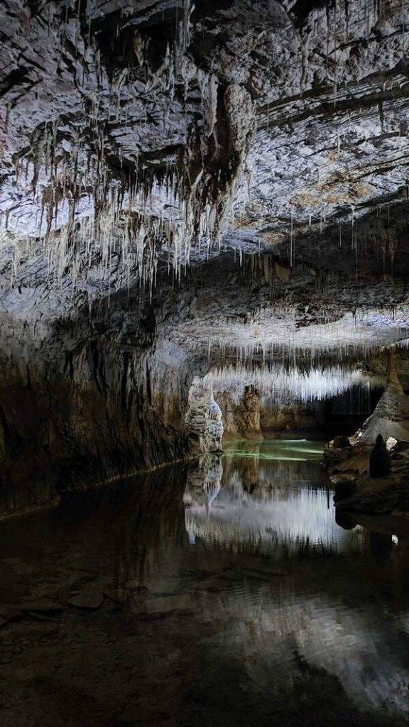 A beautifully lit cave in Choranche, France, with stalagtites trickling down above an underground river. Why do I travel? To explore the liminal places, those under the ground, or high on mountains, those which we do not normally witness.