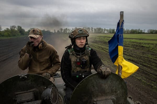 Two people on an armored vehicle with a blue and yellow flag.