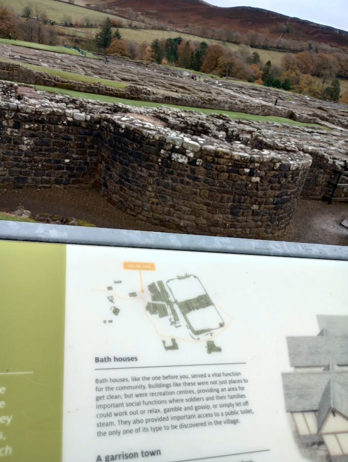 Low wall with rounded tower foundation, the remains of the bathhouse. Behind it, the remains of the Fort, and the hills