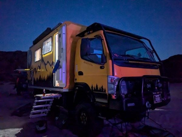 Walter the big yellow truck, against a night sky on a sandy beach with mountains silhouetted in the background