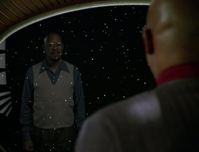Avery Brooks looking at a reflection of himself. In the foreground, his back is turned and you can see his collar and uniform. In the background, you see Benny russel dressed in a vest, collared shirt, and glasses