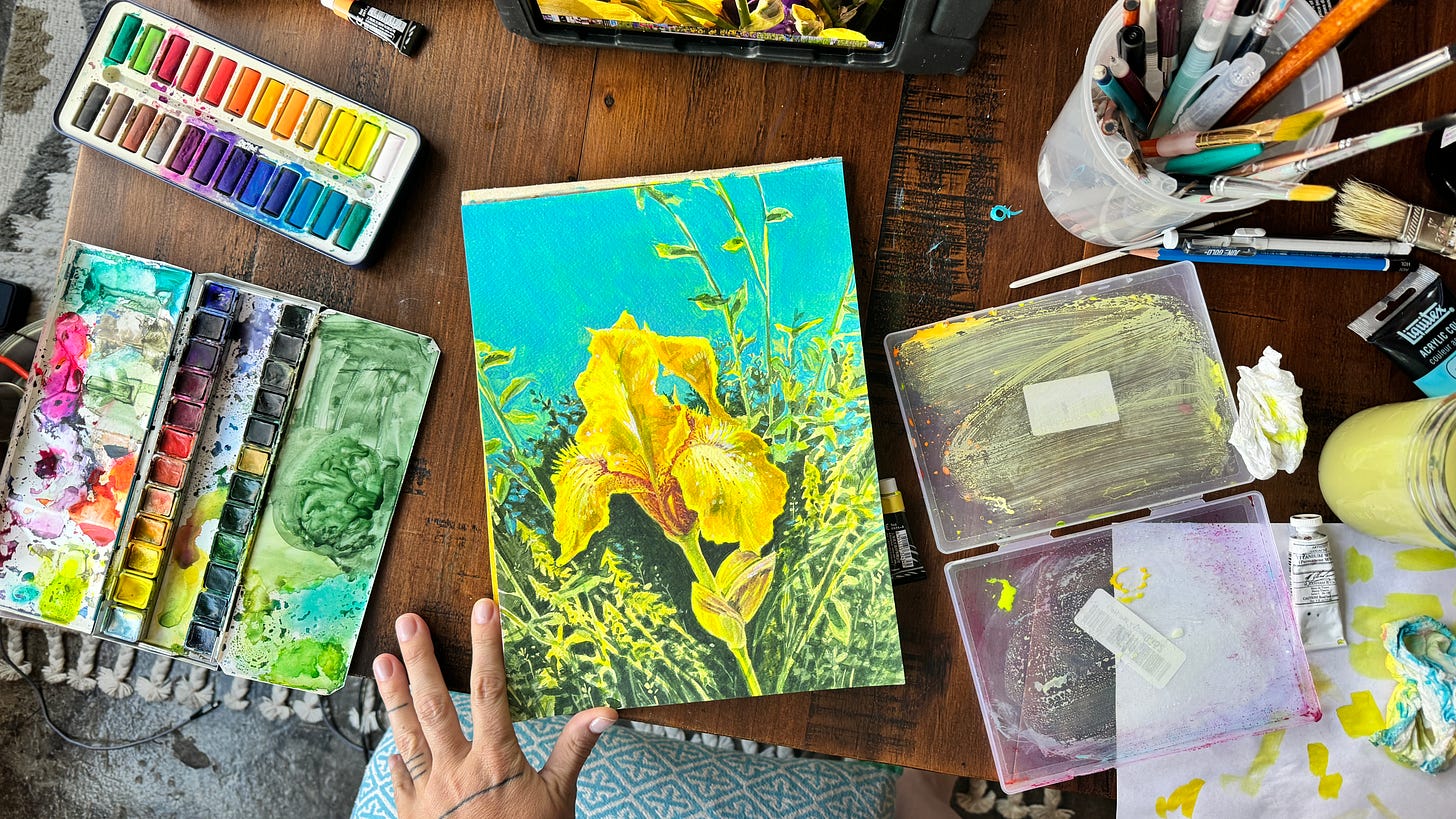 an overhead view of Lisette's watercolor setup, multiple pans of paints, brushes, and a completed painting of a yellow iris with greenery and blue sky.