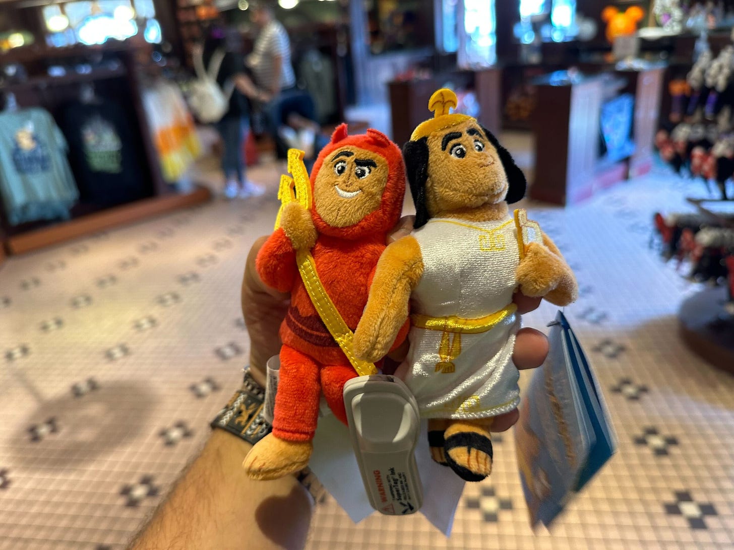 A hand holding two plush toys: one dressed as a red devil and the other as an Egyptian pharaoh. The background shows a store with various items on display, reminiscent of the enchanting shops found at Disneyland Resort.