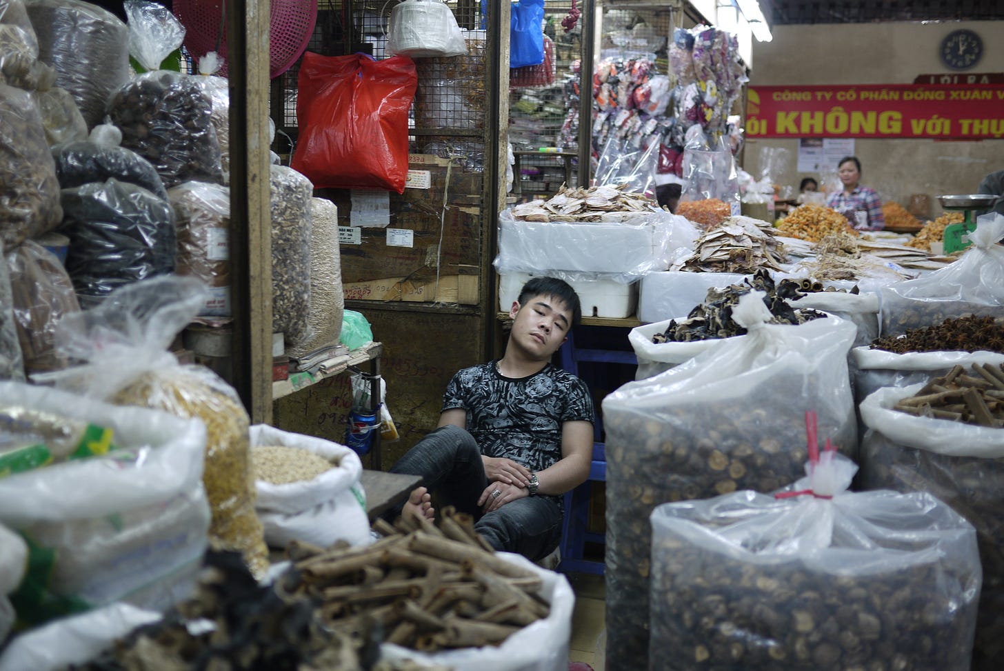 A person sleeping at a market stand between big bags of dried food.