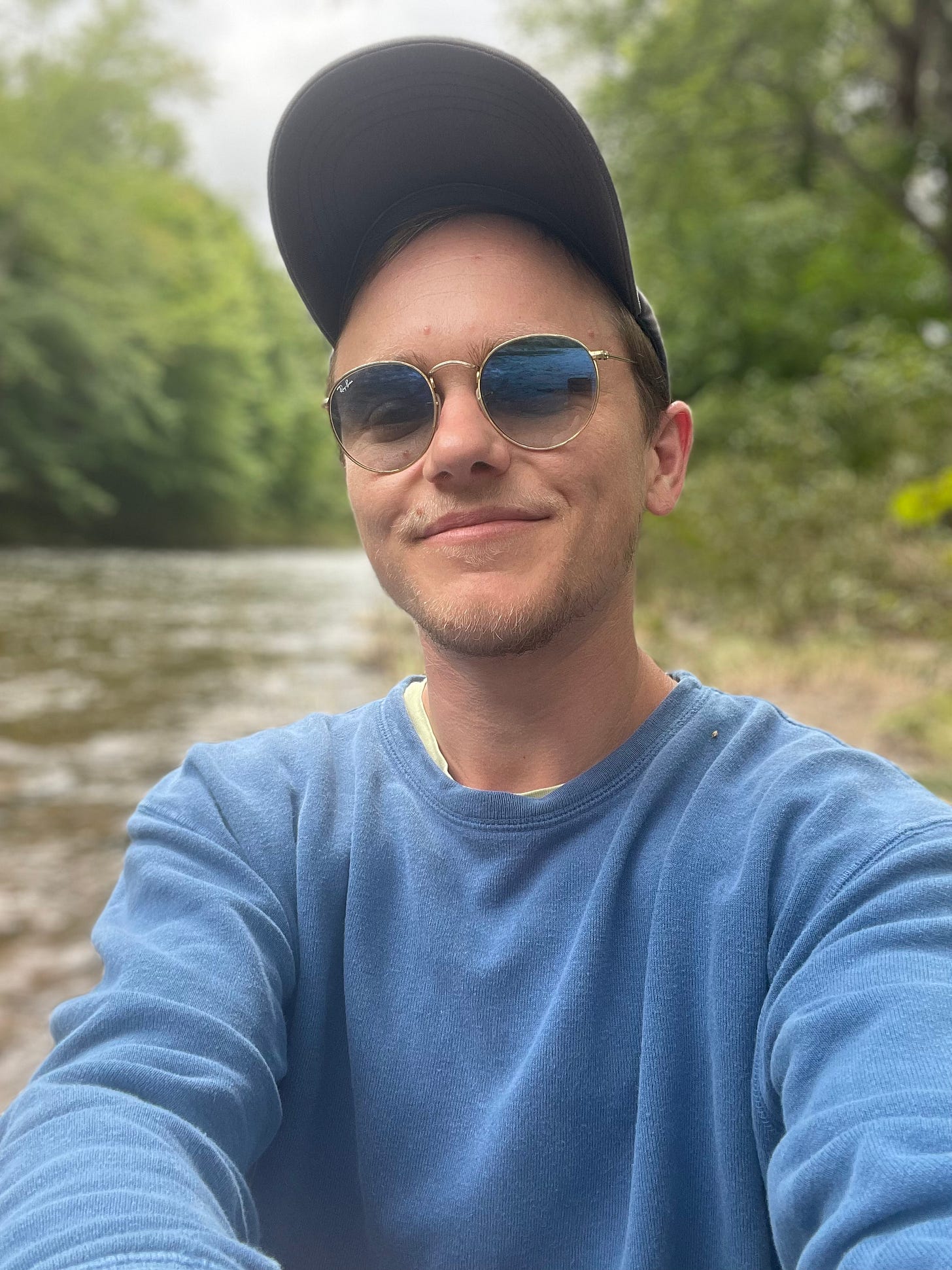 Sandy by a riverbank, wearing a blue baseball cap, sunglasses and a blue sweatshirt.