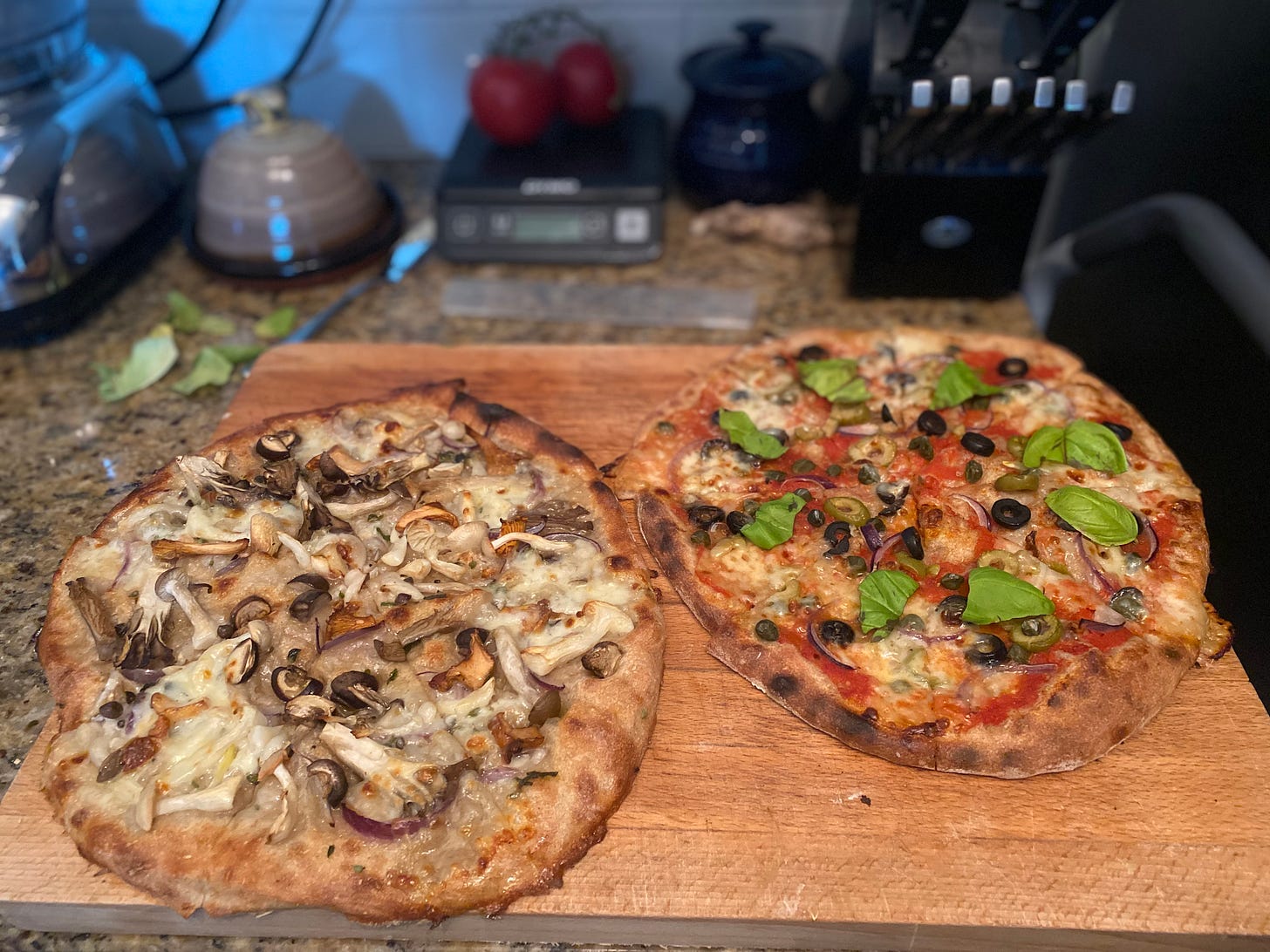 Two pizzas, as described above, on a wood cutting board. The left has big pieces of mixed mushrooms and the right has olives, capers, and large leaves of fresh basil. 