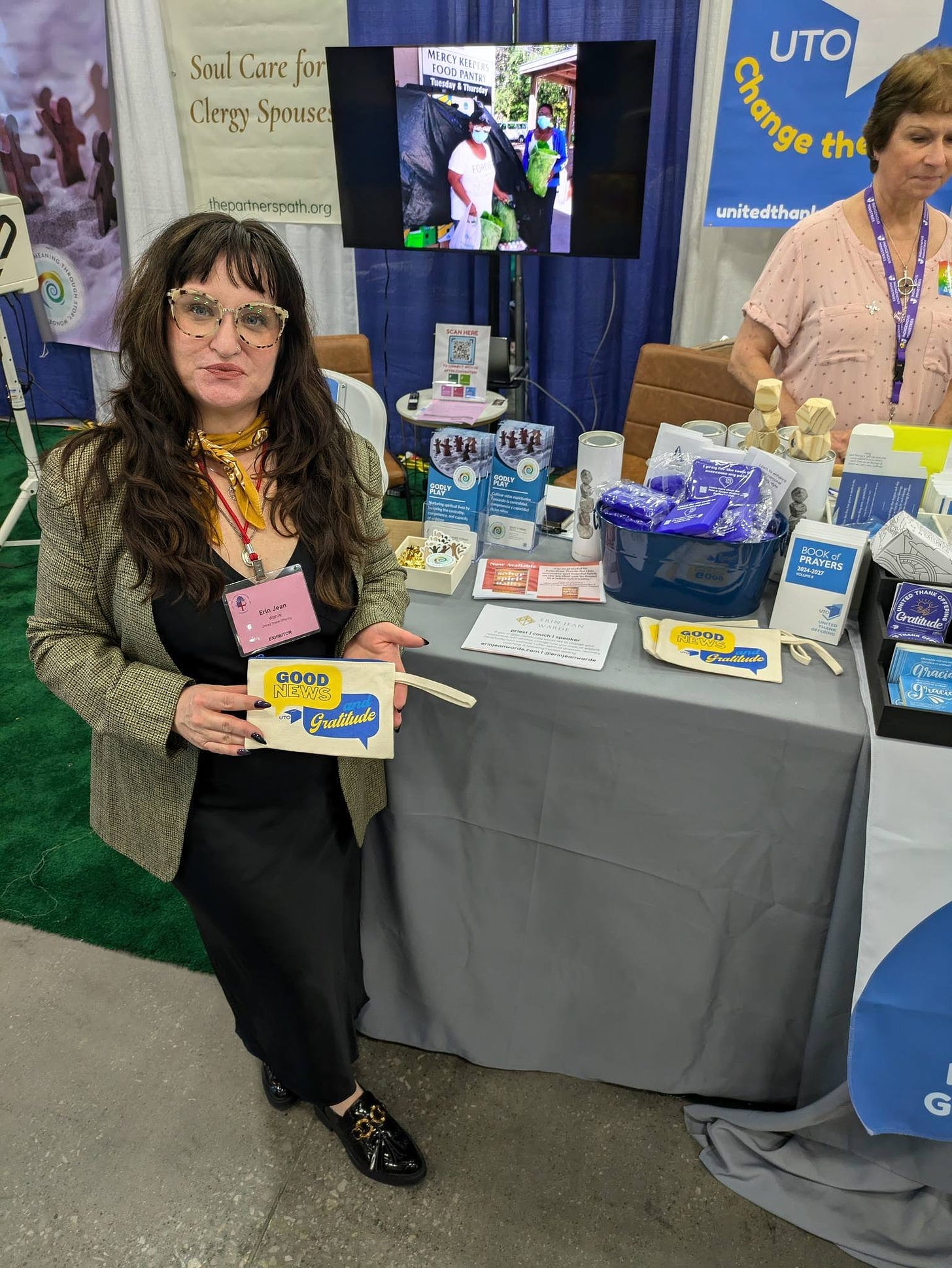 Image of EJW holding a pouch promoting the Good News & Gratitude podcast she is hosting, from UTO, at the UTO booth -- other swag is visible, other volunteers, pamphlets, etc