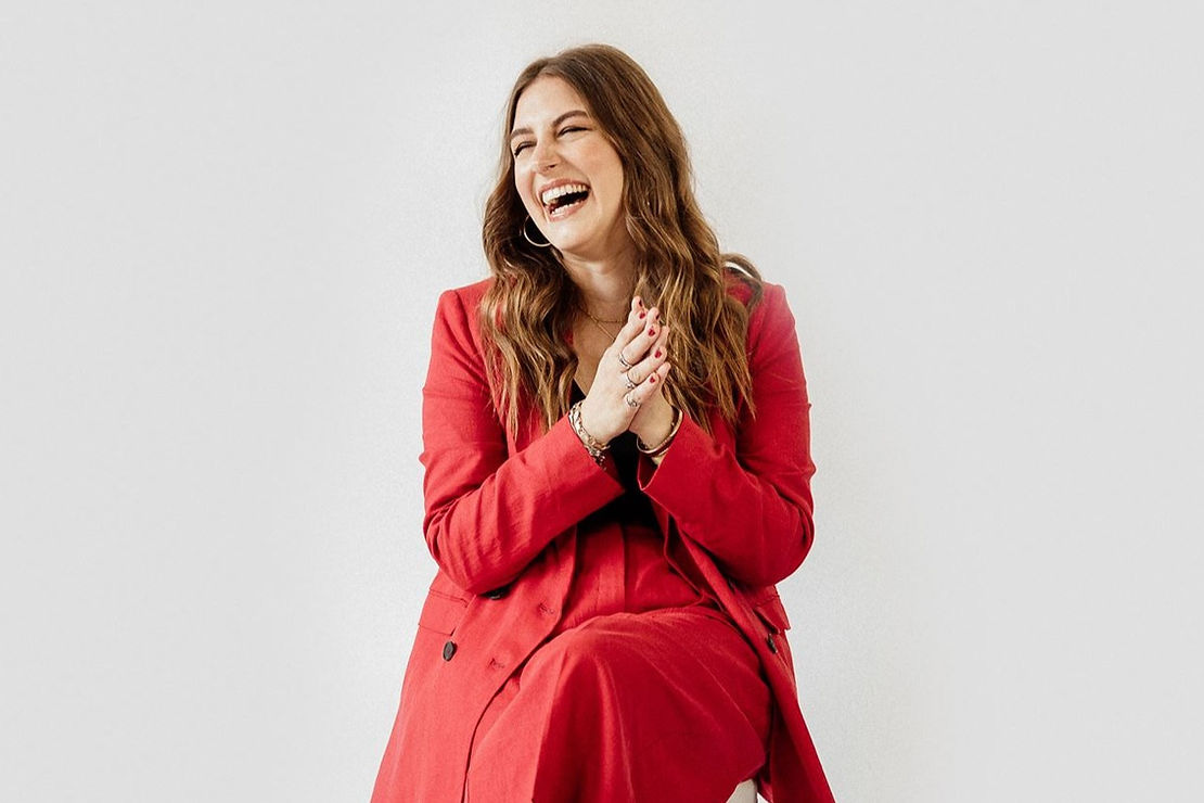 Nicole Caruso wearing a red pant suit, hands clasped and laughing.