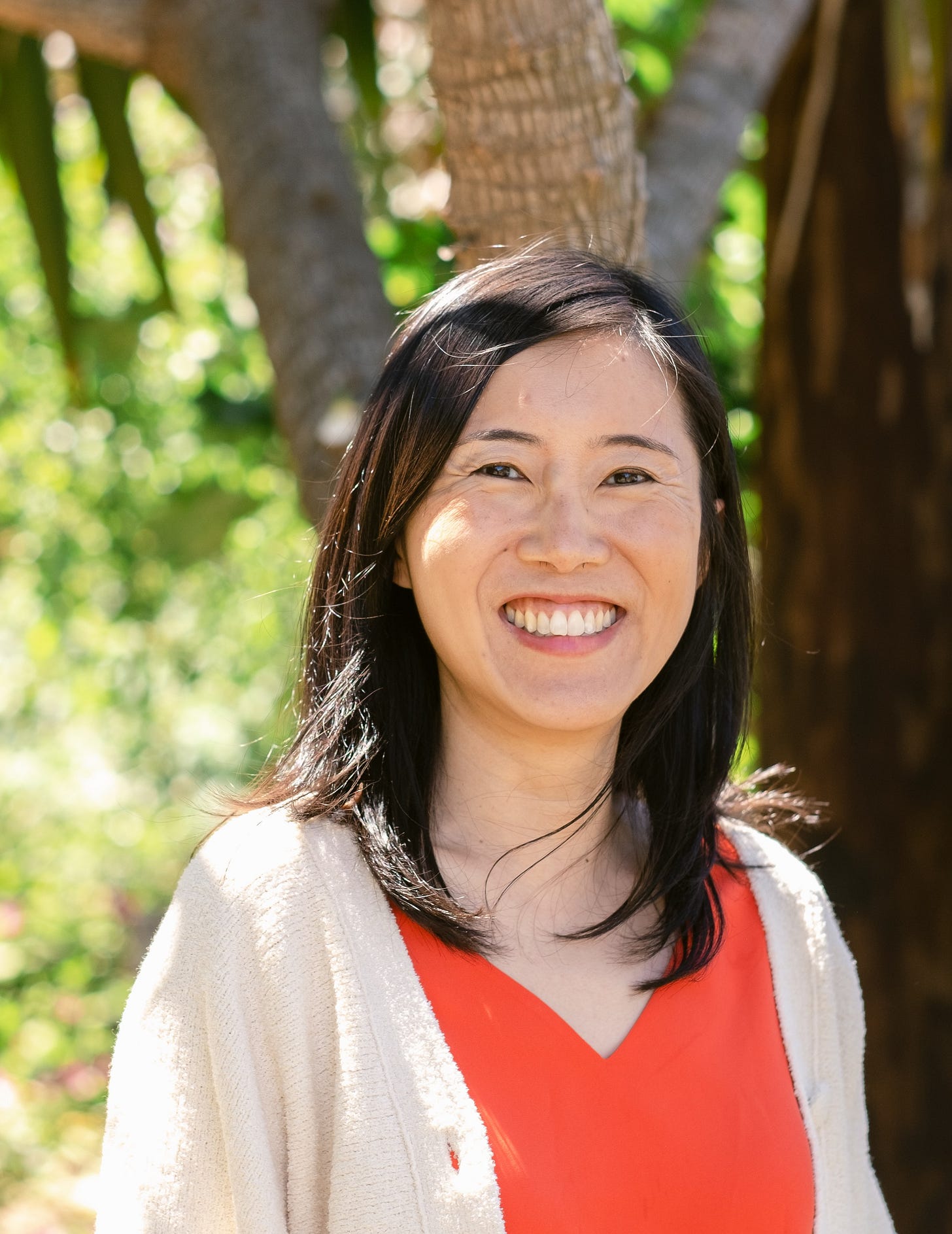 Eugenia Chiang, a Chinese woman with medium-length dark hair, smiles directly at the camera. She is outside, wearing a read shirt, and beige sweater.