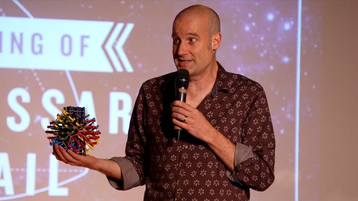 A bald man hold a microphone and a few hundred small pencils held together into the shape of a sphere by elastic bands.