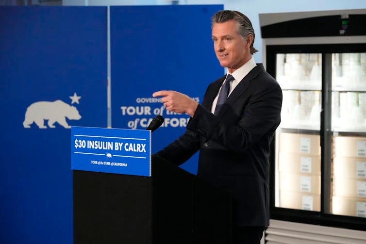 A man in a suit points in front of a lectern that says $30 insulin, with fridges of insulin in the background. A man in a suit points in front of a lectern that says $30 insulin, with fridges of insulin in the background.