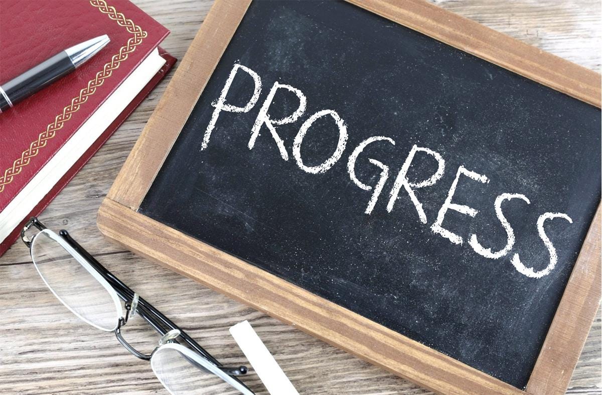 An off-kilter chalkboard with the word "progress" written on it in white all caps. To its left is a red leatherbound book with a pen on top of it, and below it are a pair of glasses and a piece of white chalk. An off-kilter chalkboard with the word "progress" written on it in white all caps. To its left is a red leatherbound book with a pen on top of it, and below it are a pair of glasses and a piece of white chalk.