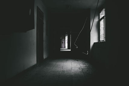 Black and white photo of a hallway in the dark with a little light shining from the window on the right and the open door in the distance.