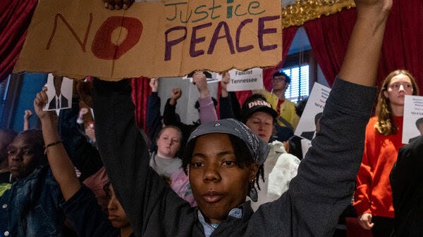 A protester holds both fists in the air, while holding on to a cardboard sign that says "No Justice No Peace." 