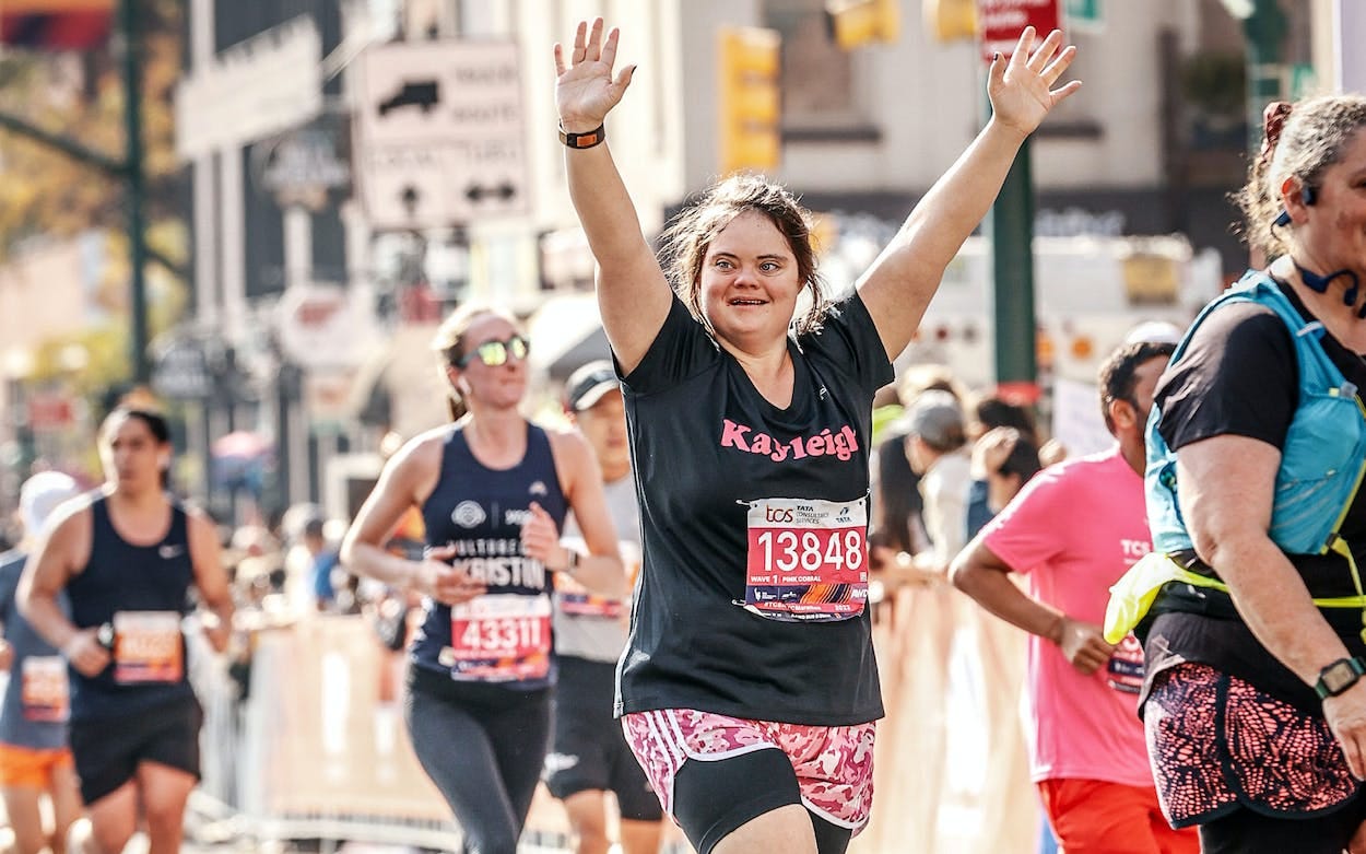 A woman with Down Syndrome holds her hands up and smiles on the running route of the NYC marathon. She's wearing a black shirt with her name, Kayleigh, in pink letters.