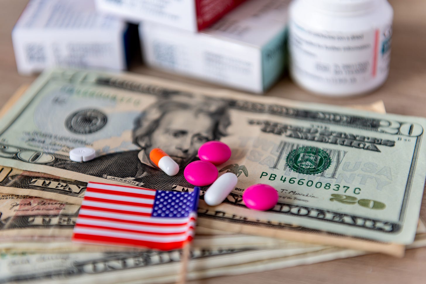 Pills on US dollar bills with a small American flag. Blurred medicine boxes in the background; focus on healthcare costs. Pills on US dollar bills with a small American flag. Blurred medicine boxes in the background; focus on healthcare costs.