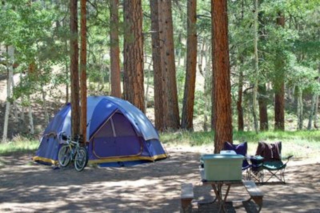 a campsite showing essential camping equipment including a blue tent, a cooler, a stove and some lawn chairs