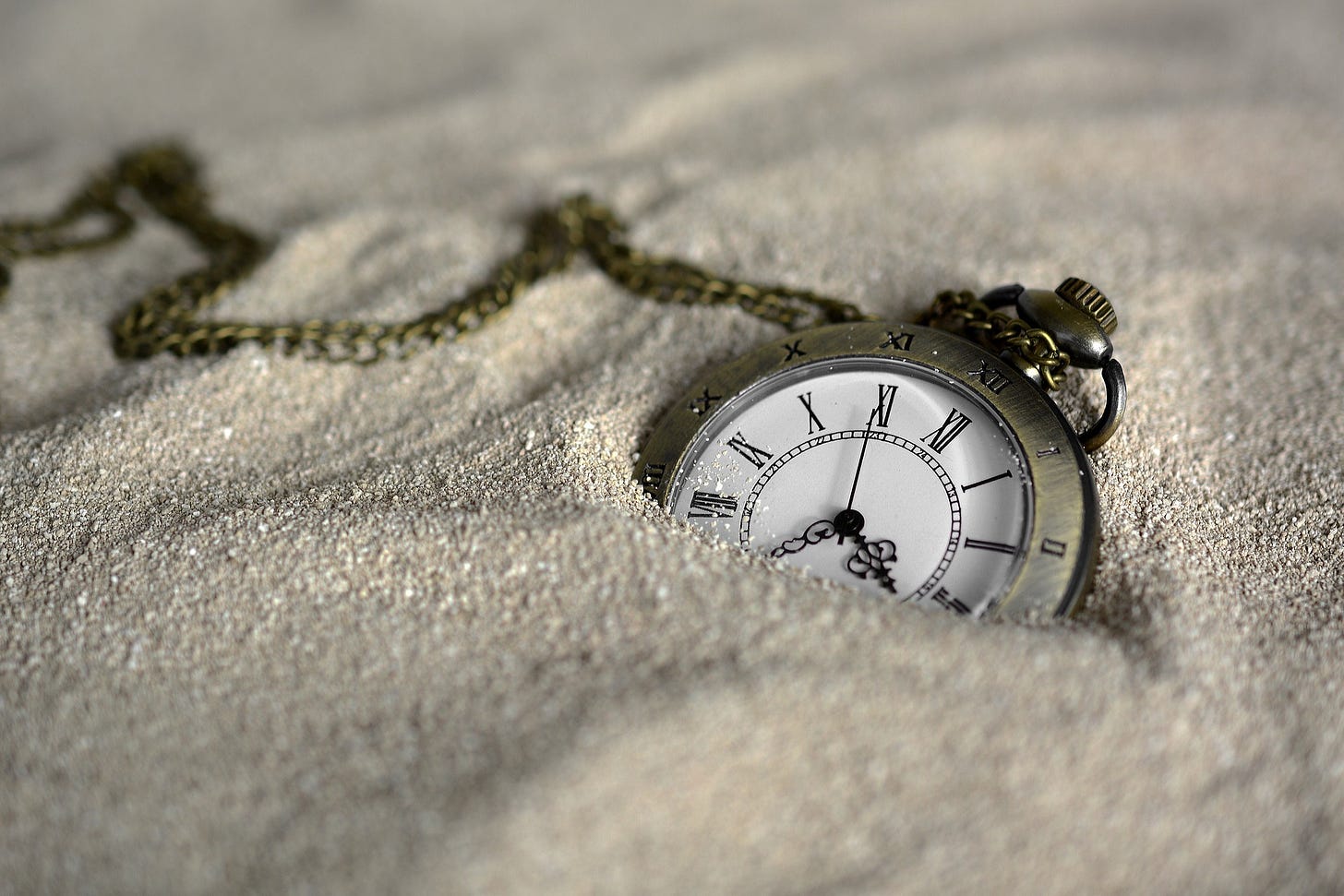 pocket watch with roman numerals and a chain half buried in silver sand