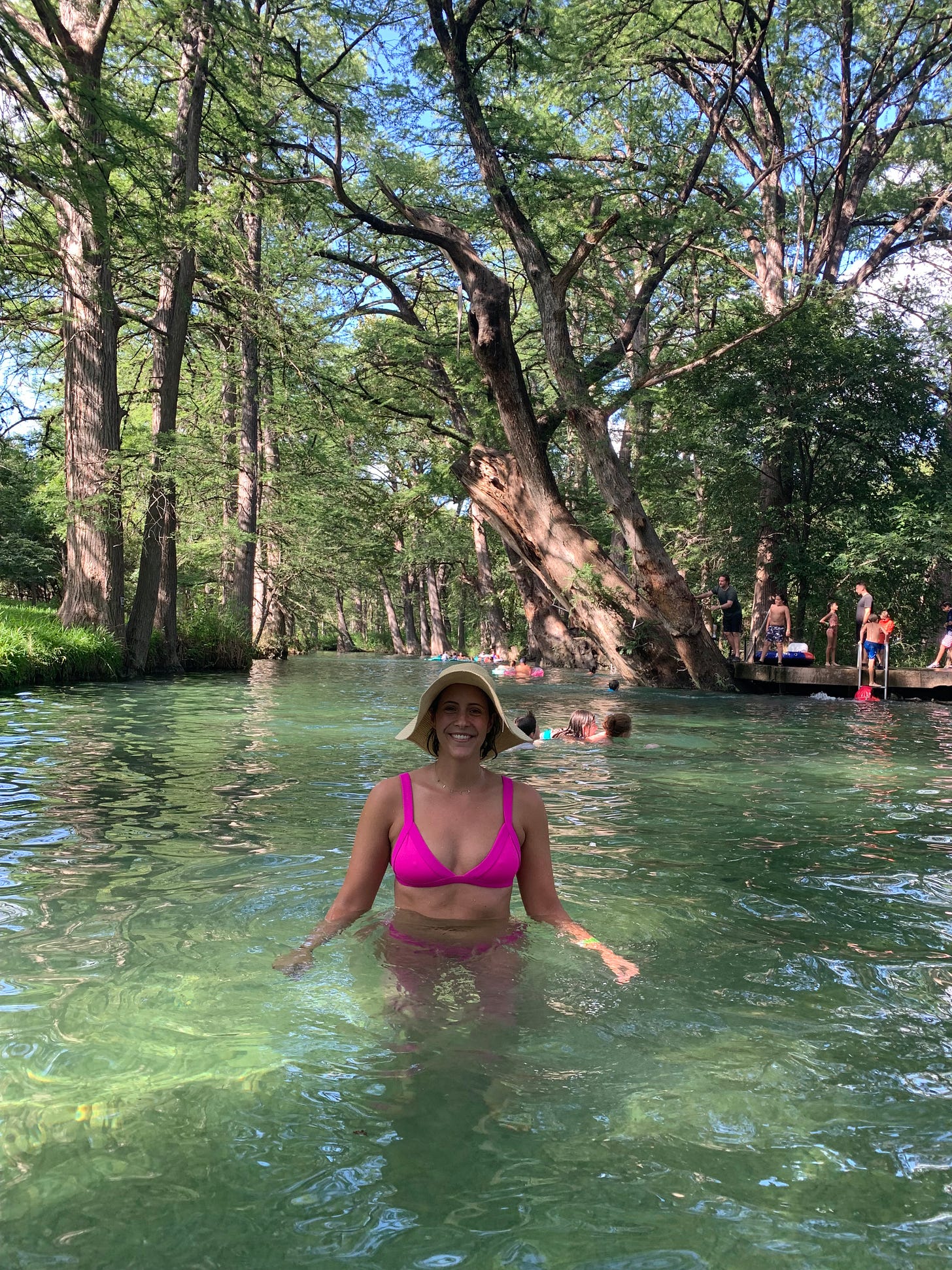 A woman smiles at the camera as she dips into spring water up to her waist. Large trees and people swimming loom in the background.
