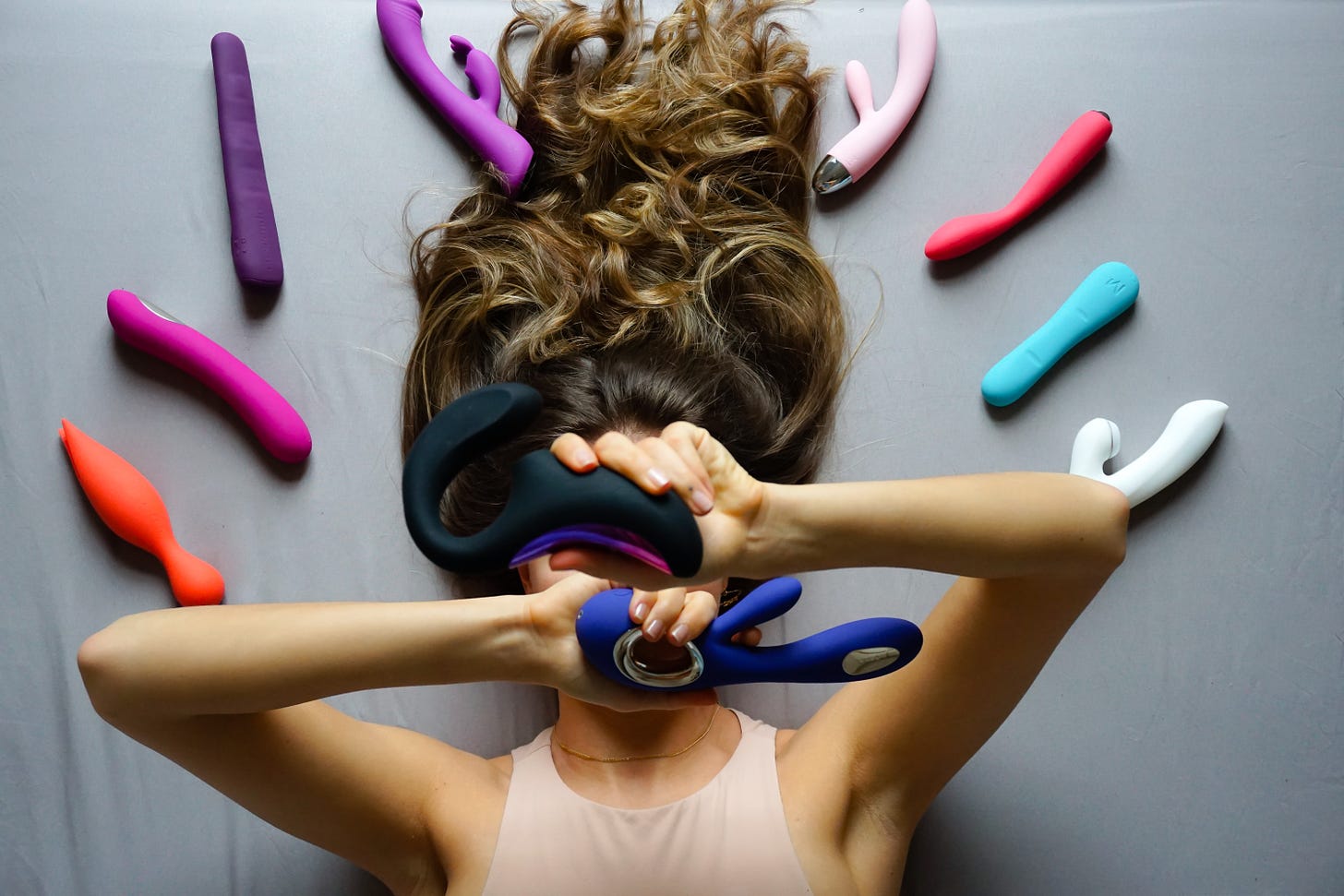 a slinder framed human with long brown hair poses with several sex toys, two hiding their face