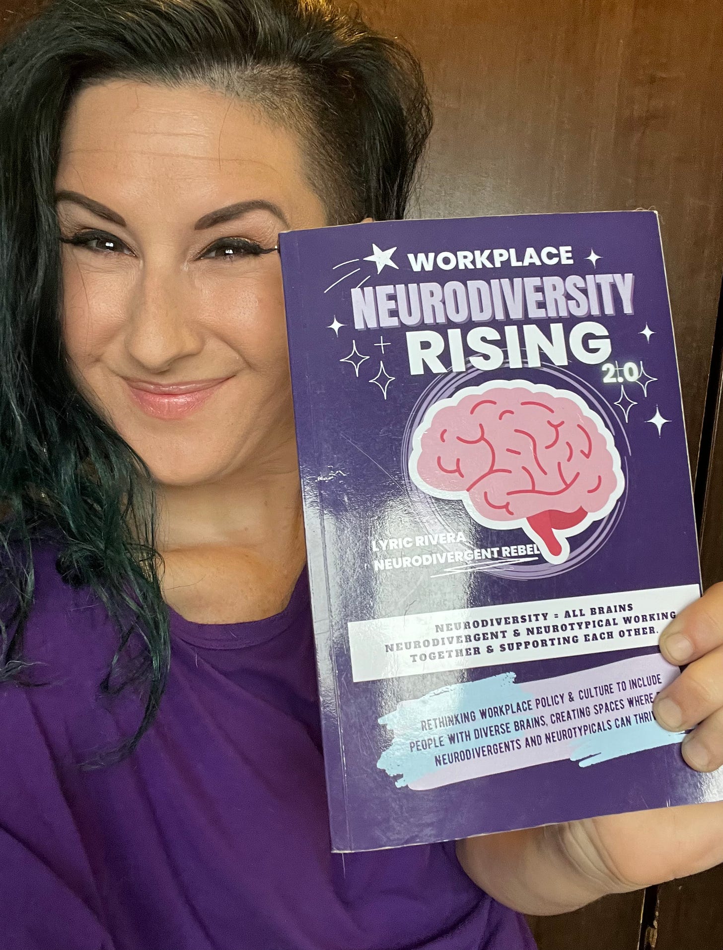 Lyric Rivera, with long green hair, winged eyeliner in a purple shirt, holding up their book “Workplace NeuroDiversity Rising” and smiling. 