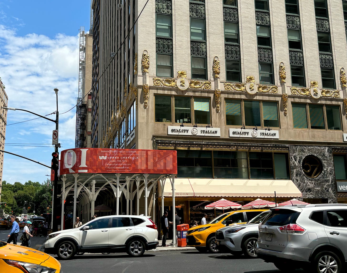 A large building on a bright day. The ground floor has signs for QUALITY ITALIAN. Over the office windows above that is gold detailing. The floors above are plainer. There is red scaffolding outside on 6th Avenue. The sky is blue and the southern end of Central Park is visible down the street.