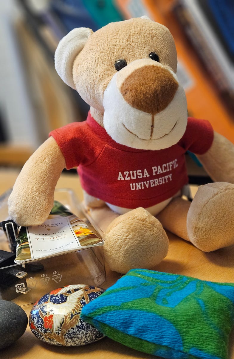 A close-up of a stuffed cougar school mascot beany baby wearing a red t-shirt with the words 'Azusa Pacific University'. Around the stuffed character are a blue and green bean bag, several palm-sized stones, hand moisturizer, hand sanitizer, and stapler remover. 